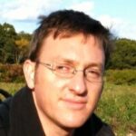 Gary Bartos poses for a candid shot on a green field with a wooden background. He has dark hair and wears glasses with a dark shirt.