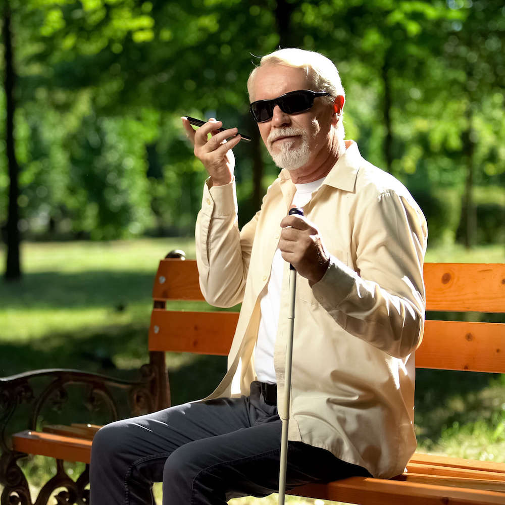 A blind man in sunglasses sits on a park bench using a voice control smartphone app