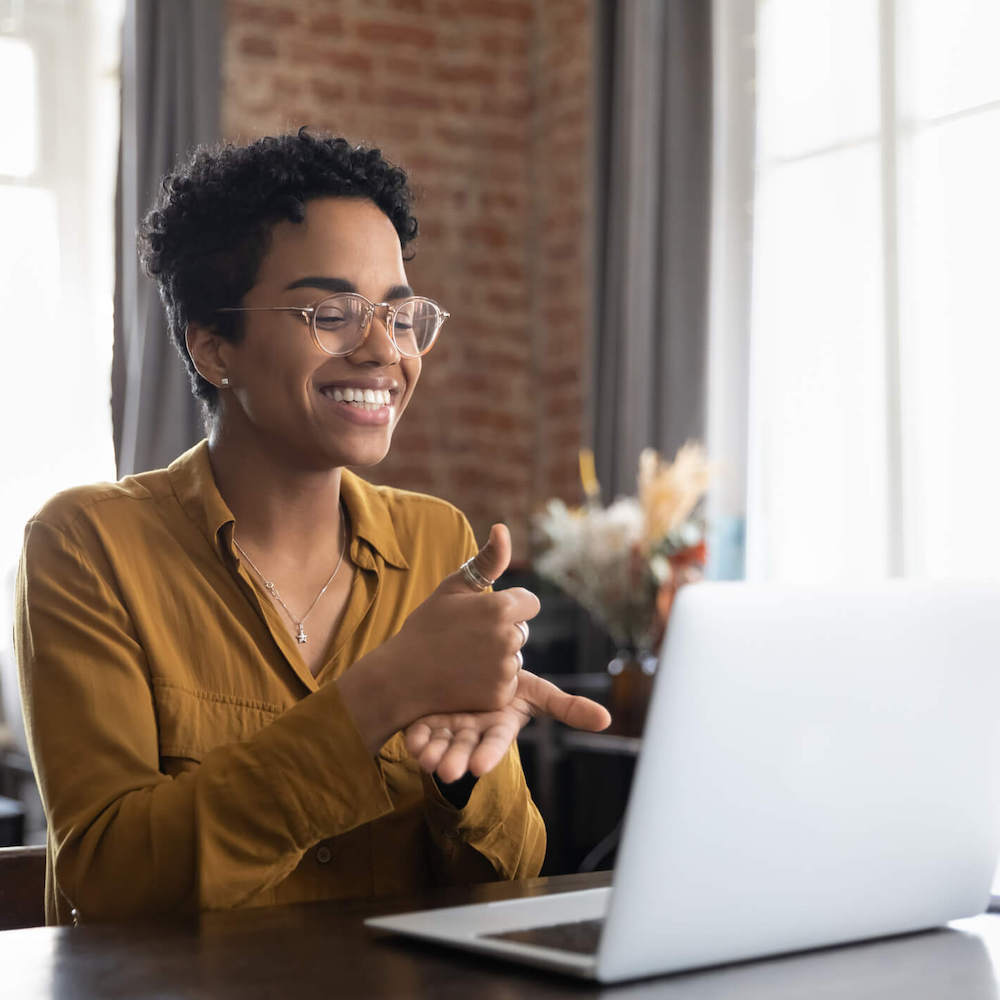 A woman smiles and makes the sign for help in front of an open laptop computer