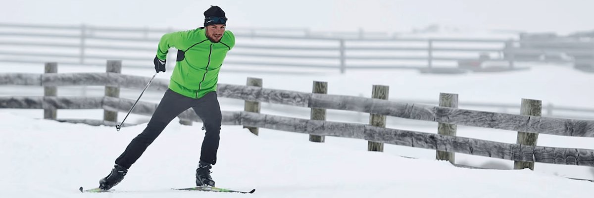 A man skate skiing through falling snow in front of a fence pushes of his right leg and ski to propel himself forward