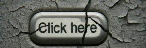 A faded and cracked "Click here" webpage button atop a gray fissured background.