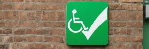 A green sign with the universal symbol of accessibility of a person in a wheelchair and a big checkmark hangs on a brick wall