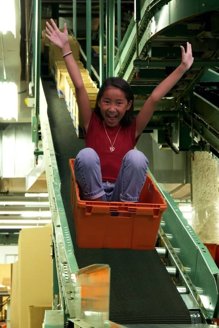 A young black girl sitting in a red tote zooms down a distribution center conveyor belt