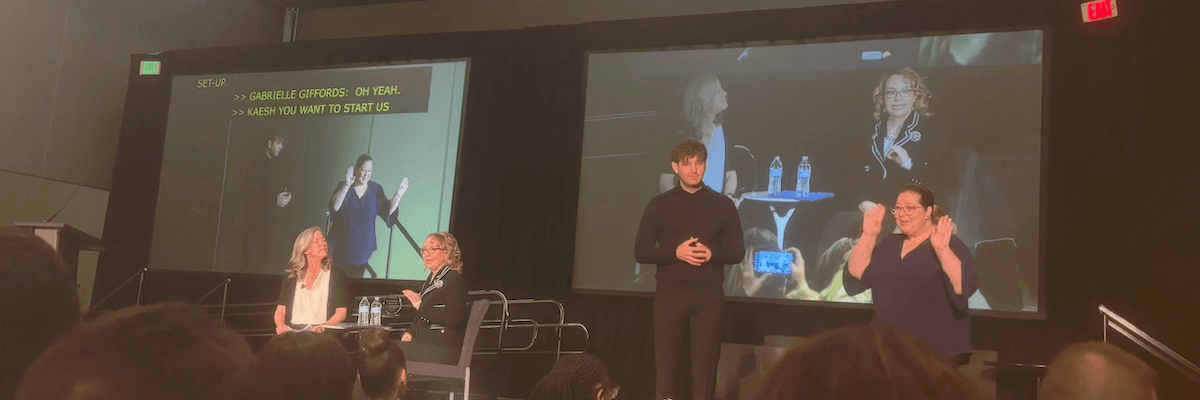 Gabby Giffords and her speech pathologist deliver the CSUN 2025 keynote speech. The pair are broadcast on a large screen while ASL interpreters and on-screen captions ensure that everyone present can understand what is being said.