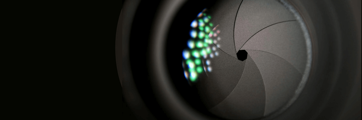 Extreme closeup of a camera shutter almost fully closed behind a lens reflecting colored lights