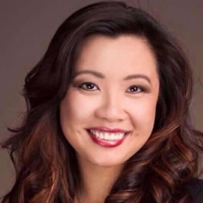 Chelsea Nguyen smiles warmly in a professional headshot. She has long, softly curled dark hair with auburn highlights, styled to one side. Her makeup is polished, with red lipstick and subtle eye shadow, and she wears a dark top against a neutral background.
