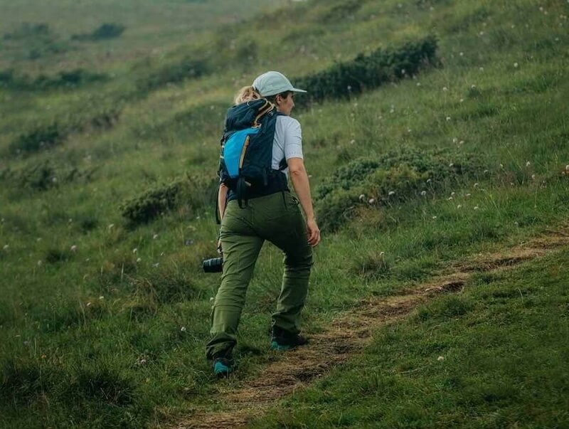 A woman wearing a baseball cap and backpack hikes up a steep, green, grassy hill.