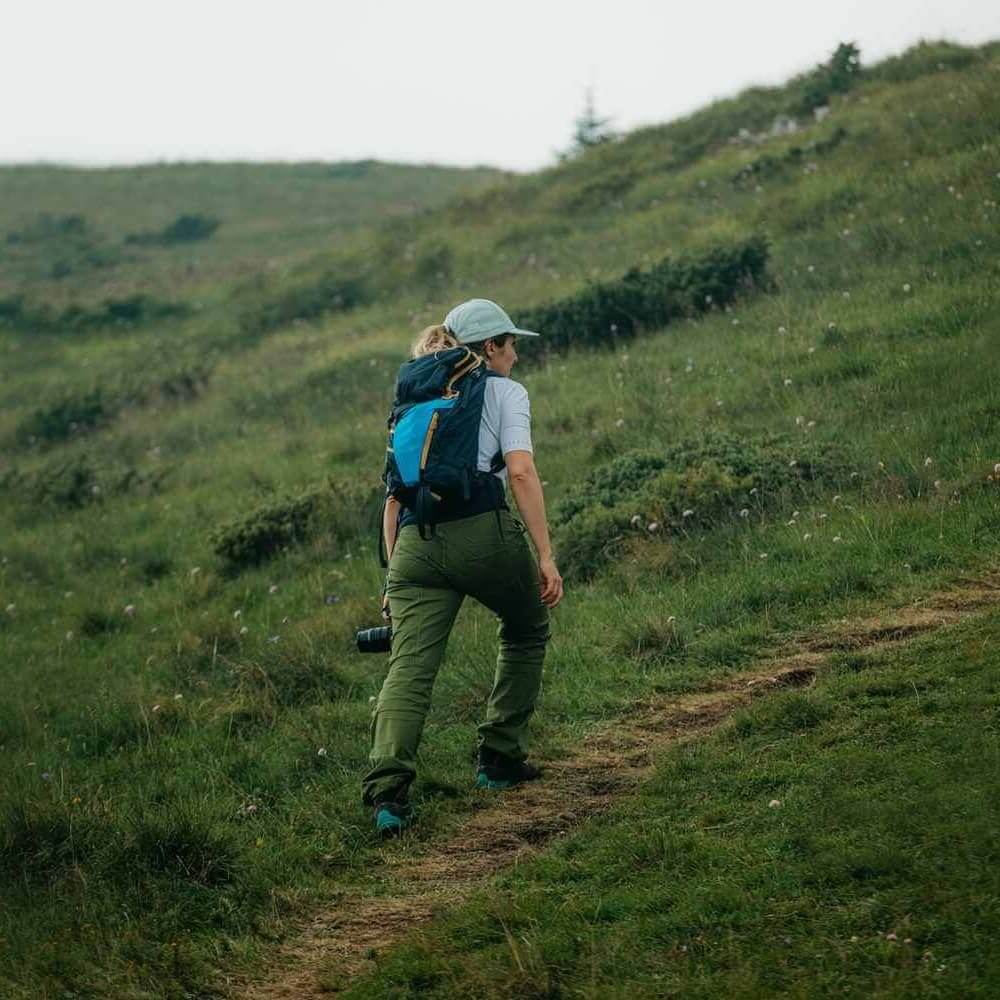 A woman wearing a baseball cap and backpack hikes up a steep, green, grassy hill.