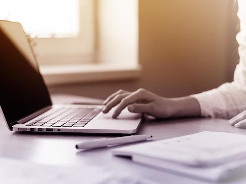 Feminine hands use a laptop on a desk with pen and paper nearby.